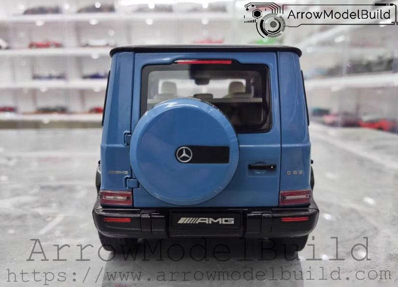 May include: A blue model car of a Mercedes-Benz G 63 AMG, viewed from the rear. The spare tire cover features the Mercedes-Benz logo. The car has black accents and the AMG logo is visible. The text "ArrowModelBuild" and a website address are at the bottom.