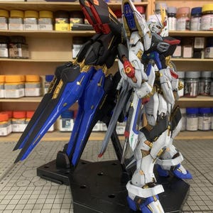 Puede incluir: Modelo detallado de un robot mecha, con detalles en blanco, azul, negro y dorado. El robot est&aacute; posado sobre una base negra, mostrando su intrincado dise&ntilde;o y estructuras similares a alas. El fondo muestra estanter&iacute;as con suministros de pintura.