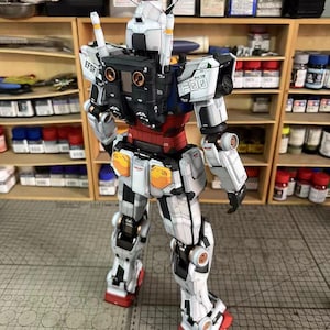 May include: A detailed model of a white, black, and red robot with orange and blue accents. The model has intricate mechanical details, including visible joints and panels. The text "EFSF" and "F03" are visible on the model. The model is on a gray surface.