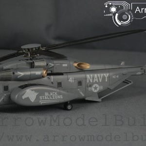 May include: A grey model helicopter with rotating blades. The words "NAVY" and "BLACK STALLIONS" are visible on the fuselage. The model has detailed features, including a tail rotor and landing gear. The text "ArrowModelBuild" is in the upper right corner.