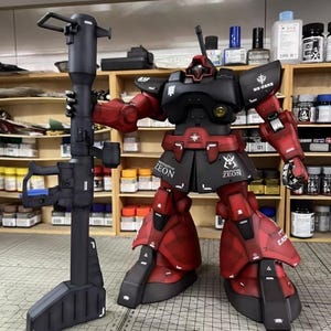 May include: A detailed model of a red and black robot figure holding a large black weapon. The robot has the text "ZEON" on its skirt and "MS-06RS" on its shoulder. The model is on a grid surface with shelves of paint and supplies in the background.