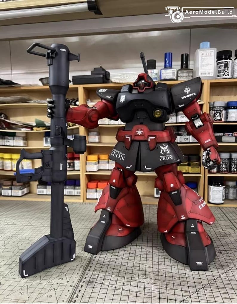 May include: A detailed model of a red and black robot figure holding a large black weapon. The robot has the text "ZEON" on its skirt and "MS-06RS" on its shoulder. The model is on a grid surface with shelves of paint and supplies in the background.