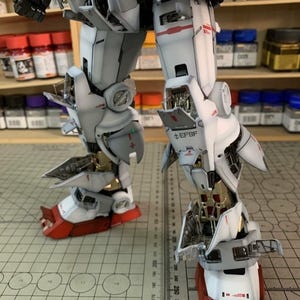 K&ouml;nnte beinhalten: Nahaufnahme der Beine eines Modellroboters, die komplexe mechanische Details zeigen. Die Beine sind &uuml;berwiegend wei&szlig; mit roten und goldenen Akzenten. Der Text "EFSF" ist sichtbar. Das Modell befindet sich auf einer Schneidematte.