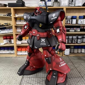 May include: A detailed model of a red and black robot with intricate detailing. The robot has a black head and torso, with red arms, legs, and feet. The model has the word "ZEON" printed on the skirt.
