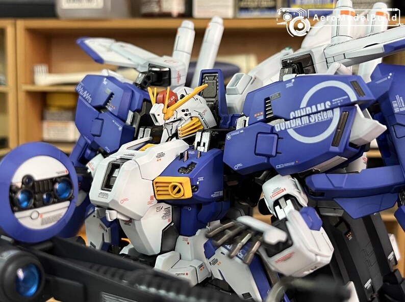 May include: A detailed model of a Gundam robot, mainly blue, white, and yellow. The model has intricate mechanical details, including panels, vents, and articulated joints. The text "GUNDAM system" is visible on the shoulder. A collectible item.