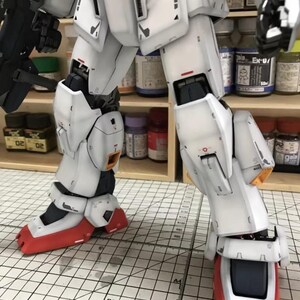 Op de afbeelding: Een gedetailleerd, gedeeltelijk geassembleerd wit en grijs model robot met rode en zwarte accenten. Het model heeft ingewikkelde paneellijnen en mechanische details, met een zichtbaar pistool. Het model staat op een rasteroppervlak, met verfpotten op de achtergrond.