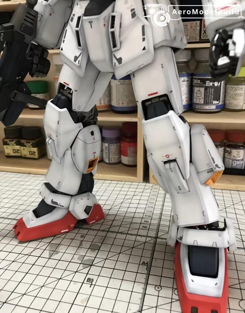Op de afbeelding: Een gedetailleerd, gedeeltelijk geassembleerd wit en grijs model robot met rode en zwarte accenten. Het model heeft ingewikkelde paneellijnen en mechanische details, met een zichtbaar pistool. Het model staat op een rasteroppervlak, met verfpotten op de achtergrond.