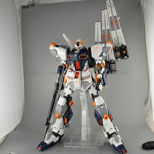 May include: A detailed model of a white, navy, and orange mecha robot with large shoulder-mounted weapons. The robot is standing on a clear plastic stand. The model has intricate details and is posed in a dynamic stance.
