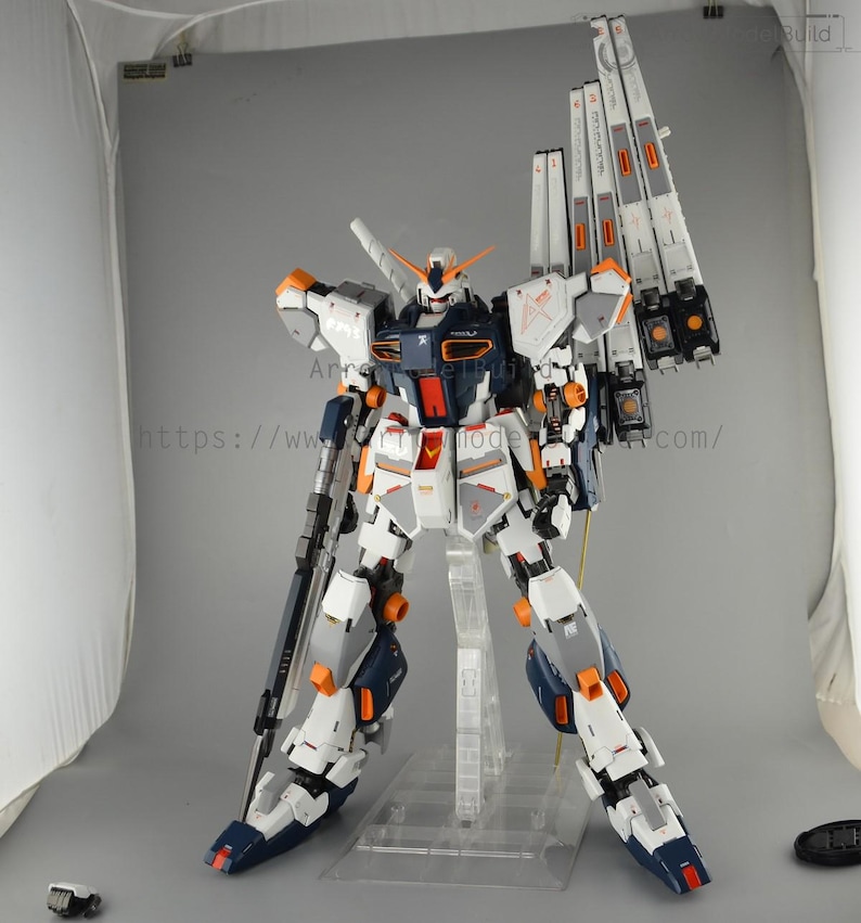 May include: A detailed model of a white, navy, and orange mecha robot with large shoulder-mounted weapons. The robot is standing on a clear plastic stand. The model has intricate details and is posed in a dynamic stance.