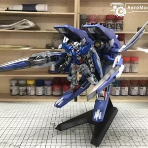 May include: A detailed model of a blue and white mecha robot, mounted on a black stand. The robot has intricate details and is posed with a large weapon. The background shows shelves with paint bottles and tools, suggesting a hobbyist's workspace.