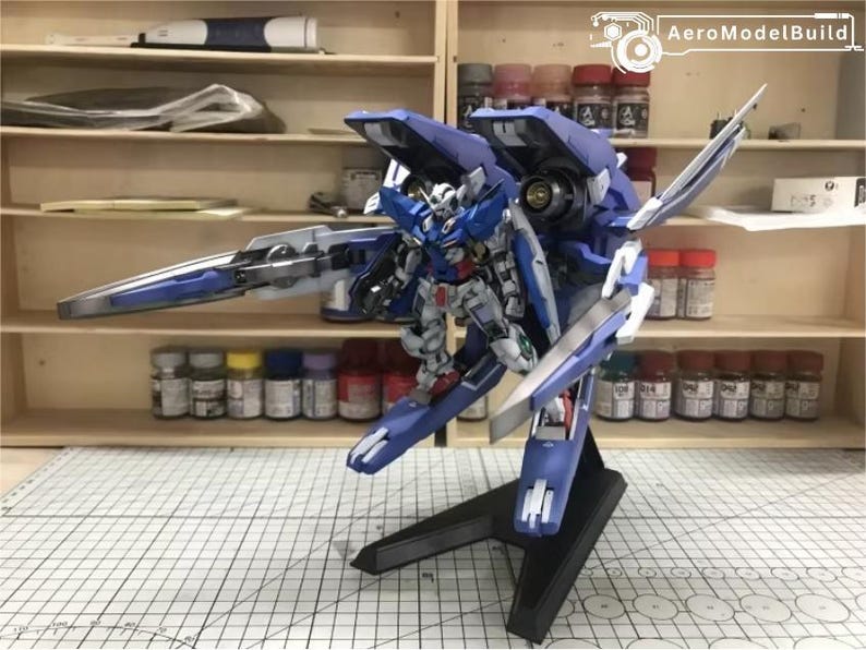 May include: A detailed model of a blue and white mecha robot, mounted on a black stand. The robot has intricate details and is posed with a large weapon. The background shows shelves with paint bottles and tools, suggesting a hobbyist's workspace.