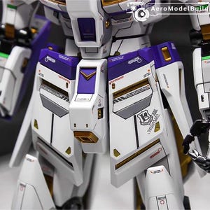 May include: Close-up of a detailed white and purple model robot. The model features gold accents, gray panels, and various markings. The design includes sharp angles and geometric shapes, with the text "AeroModelBuild" visible.