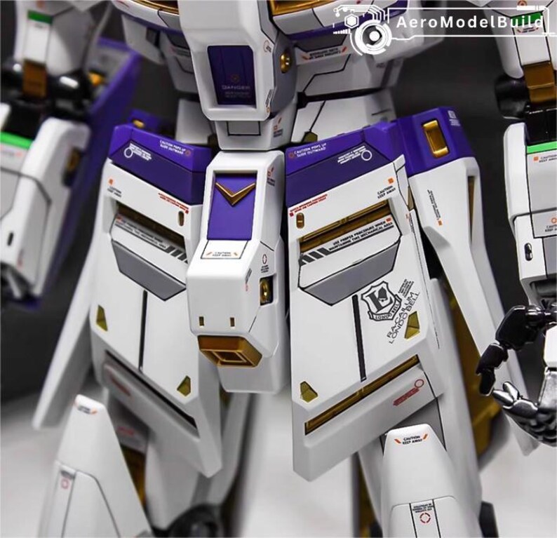 May include: Close-up of a detailed white and purple model robot. The model features gold accents, gray panels, and various markings. The design includes sharp angles and geometric shapes, with the text "AeroModelBuild" visible.