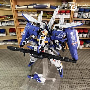May include: A detailed model of a blue, white, and yellow mecha robot with a large black rifle. The robot has multiple wings and panels, and is mounted on a clear stand. The background includes shelves with paint bottles.
