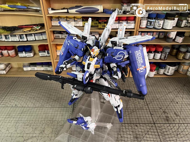 May include: A detailed model of a blue, white, and yellow mecha robot with a large black rifle. The robot has multiple wings and panels, and is mounted on a clear stand. The background includes shelves with paint bottles.