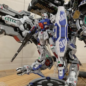 May include: A detailed model of a Gundam robot, primarily white, blue, and red, with intricate mechanical details. The robot is equipped with a shield and a large weapon, standing on a circular base. The model is a complex assembly, showcasing various components.