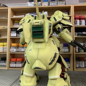 May include: A detailed, light yellow model kit of a mobile suit, featuring intricate mechanical details. The model has a large backpack with green vents, a long antenna, and a black weapon. The text "ArrowModelBuild" is visible.
