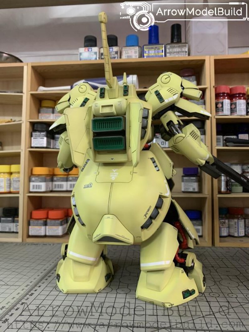 May include: A detailed, light yellow model kit of a mobile suit, featuring intricate mechanical details. The model has a large backpack with green vents, a long antenna, and a black weapon. The text "ArrowModelBuild" is visible.