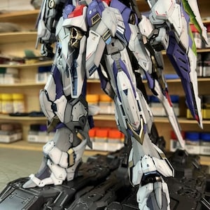 May include: A highly detailed robot model, predominantly white, grey, purple and red. The model is posed on a black surface, highlighting its intricate mechanical design. The model's design includes sharp angles and complex paneling.