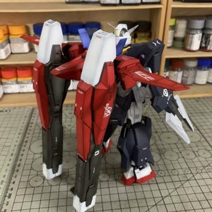 Puede incluir: Un modelo detallado de un robot mec&aacute;nico, con una combinaci&oacute;n de colores rojo, blanco, negro y azul. El modelo tiene grandes piezas de armadura angulares y alas. El modelo est&aacute; sobre una esterilla de corte con una regla.