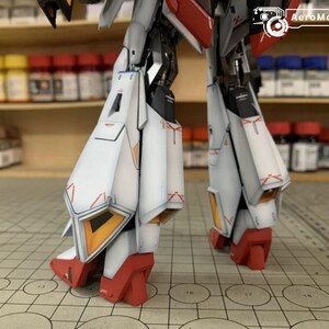 May include: Close-up of a detailed model robot's legs. The legs are primarily white with red and orange accents. The model is on a grid surface, with shelves of paint bottles in the background. The AeroModelBuild logo is visible.