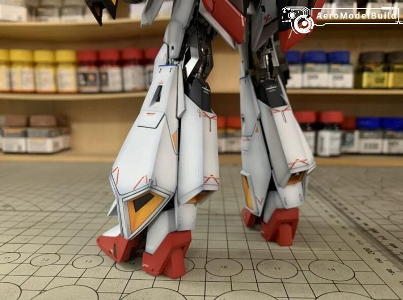 May include: Close-up of a detailed model robot's legs. The legs are primarily white with red and orange accents. The model is on a grid surface, with shelves of paint bottles in the background. The AeroModelBuild logo is visible.