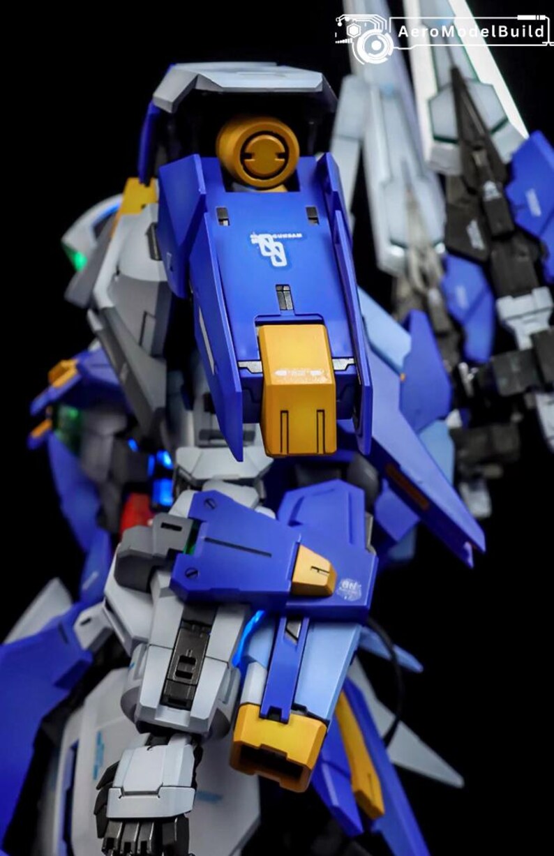 May include: Close-up of a detailed blue, gray, and yellow model robot. The robot has intricate mechanical details, with visible joints, panels, and accents. The model is set against a dark background, highlighting its design.