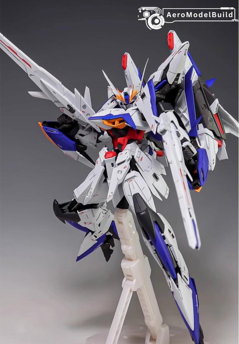 Puede incluir: Maqueta detallada de un robot futurista, predominantemente blanco con detalles azules, rojos y negros. El robot presenta un intrincado blindaje, grandes alas y una cabeza estilizada. Est&aacute; montado sobre un soporte blanco.