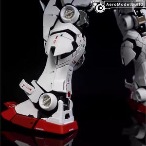 Op de afbeelding: Close-up van een wit en rood model robotbeen. De poot heeft gedetailleerde panelen, rode accenten op de voet en kleine groene en rode markeringen. De tekst "AeroModelBuild" is zichtbaar in de rechterbovenhoek.