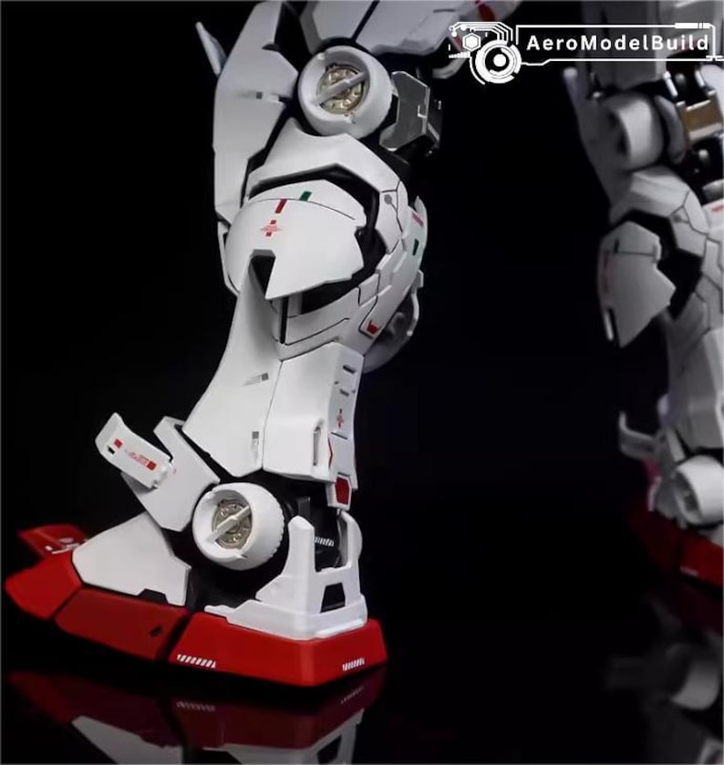 Op de afbeelding: Close-up van een wit en rood model robotbeen. De poot heeft gedetailleerde panelen, rode accenten op de voet en kleine groene en rode markeringen. De tekst "AeroModelBuild" is zichtbaar in de rechterbovenhoek.