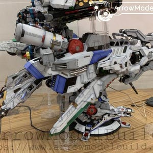 May include: A detailed model of a futuristic robot, primarily white with blue, red, and green accents. The model features intricate mechanical details, including multiple articulated parts and weapons. The model is displayed on a wooden surface.