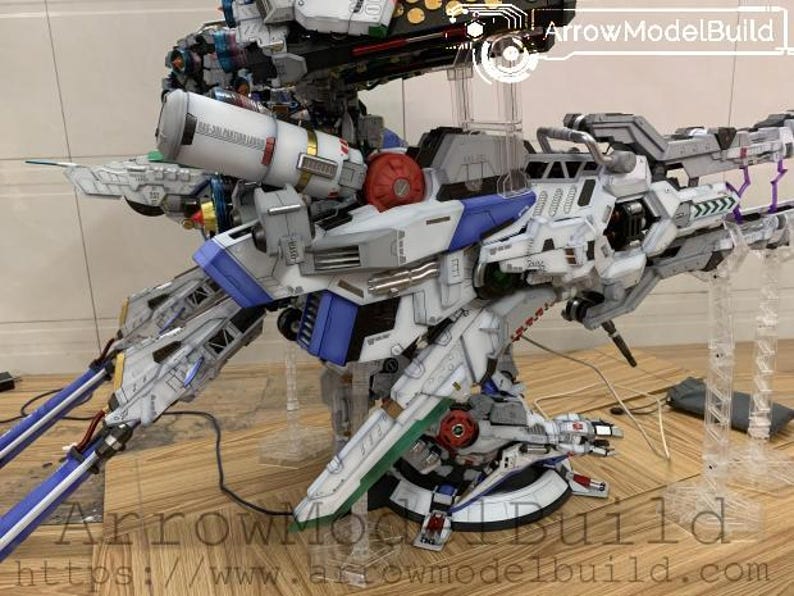 May include: A detailed model of a futuristic robot, primarily white with blue, red, and green accents. The model features intricate mechanical details, including multiple articulated parts and weapons. The model is displayed on a wooden surface.