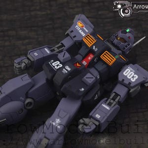 Puede incluir: Modelo detallado de un robot mecánico morado y negro, tumbado sobre una superficie de madera. El modelo tiene detalles en naranja y blanco, con la palabra "TITANS" visible. El número "03" está impreso en el modelo.