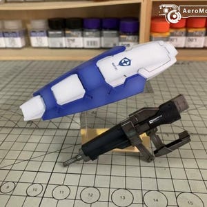 Puede incluir: Un componente de kit de modelo azul y blanco con forma de escudo, con un logotipo. Tambi&eacute;n hay una pistola modelo negra y plateada. Los art&iacute;culos est&aacute;n sobre una superficie de cuadr&iacute;cula, con botes de pintura en el fondo.