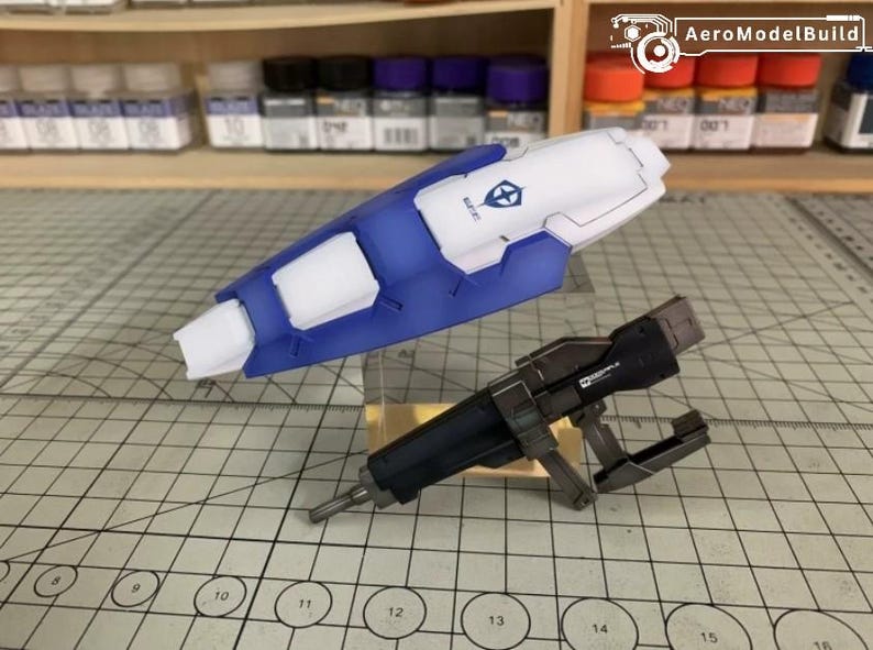 Puede incluir: Un componente de kit de modelo azul y blanco con forma de escudo, con un logotipo. Tambi&eacute;n hay una pistola modelo negra y plateada. Los art&iacute;culos est&aacute;n sobre una superficie de cuadr&iacute;cula, con botes de pintura en el fondo.