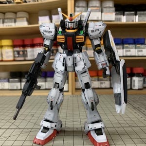 Pode incluir: Modelo detalhado de um Gundam RX-178, predominantemente branco com detalhes cinza e preto. O modelo apresenta um rifle preto, um escudo e p&eacute;s vermelhos e brancos. Est&aacute; posicionado sobre uma superf&iacute;cie quadriculada, com uma prateleira de potes de tinta ao fundo.