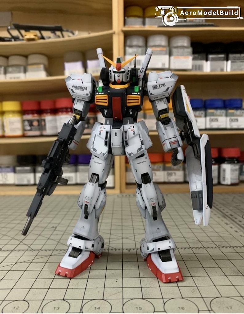Pode incluir: Modelo detalhado de um Gundam RX-178, predominantemente branco com detalhes cinza e preto. O modelo apresenta um rifle preto, um escudo e p&eacute;s vermelhos e brancos. Est&aacute; posicionado sobre uma superf&iacute;cie quadriculada, com uma prateleira de potes de tinta ao fundo.