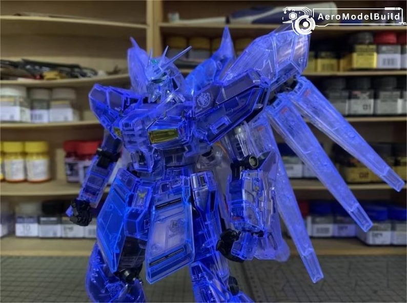 May include: A detailed, translucent blue model robot, featuring articulated limbs and wing-like elements. The robot is displayed in a dynamic pose, highlighting its intricate design. Shelves with containers are in the background, indicating a hobby or model-making setting. The text "AeroModelBuild" is visible.