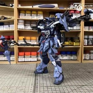 May include: A detailed model of a blue and gray mech robot, with intricate armor and weaponry. The model is posed on a grid surface, with shelves of paint and supplies in the background. AeroModelBuild logo is visible.