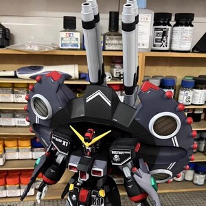 May include: A detailed model of a black, gray, and red mecha robot with large shoulder armor and multiple missile launchers. The model has yellow and red accents, with the text "GFFS-X1" and "DESTROY" visible. The model is on a shelf with various model-building supplies.
