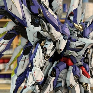 May include: A detailed model robot with a futuristic design, featuring white, purple, and gold accents. The model has intricate mechanical details, suggesting it is a collectible item or a hobby project.