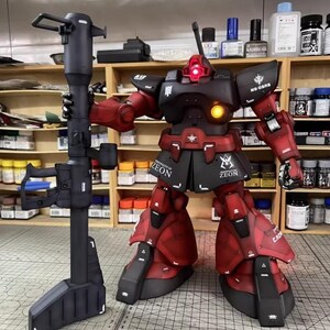 May include: A detailed model of a red and black robot holding a large black weapon. The robot has the word "ZEON" printed on its skirt and is posed in a workshop setting with paint bottles and shelves in the background.
