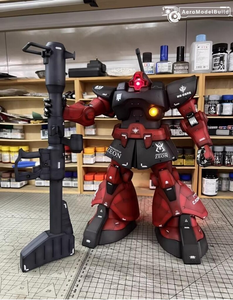May include: A detailed model of a red and black robot holding a large black weapon. The robot has the word "ZEON" printed on its skirt and is posed in a workshop setting with paint bottles and shelves in the background.