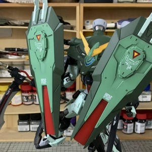 Puede incluir: Modelo detallado de un robot mecha verde y rojo con grandes estructuras en forma de escudo. El robot tiene detalles intrincados, incluyendo acentos amarillos y un dise&ntilde;o complejo. El modelo se encuentra sobre un fondo de estanter&iacute;as con diversas herramientas y suministros.