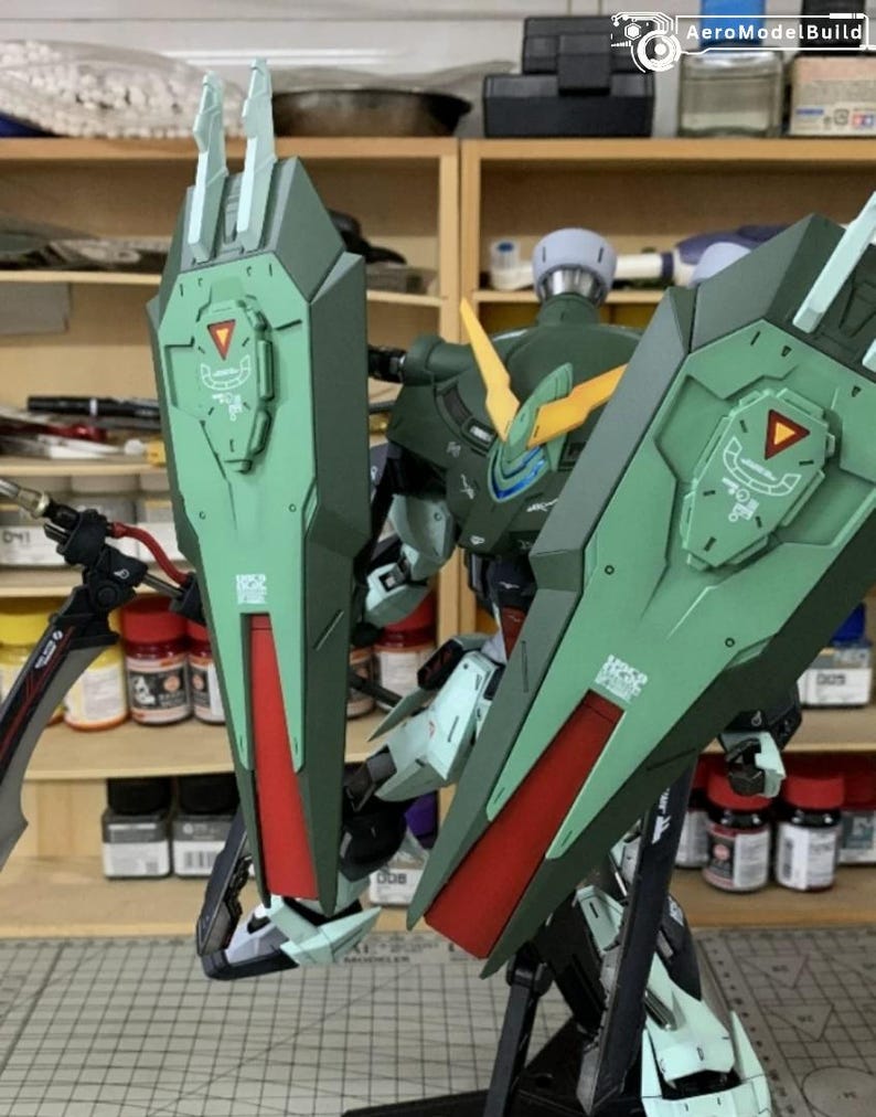 Puede incluir: Modelo detallado de un robot mecha verde y rojo con grandes estructuras en forma de escudo. El robot tiene detalles intrincados, incluyendo acentos amarillos y un dise&ntilde;o complejo. El modelo se encuentra sobre un fondo de estanter&iacute;as con diversas herramientas y suministros.