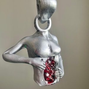 May include: A silver pendant necklace depicting a stylized torso with an open chest cavity filled with red, organic details. The pendant hangs from a silver chain.