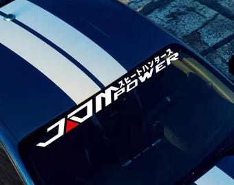 Pode incluir: Uma faixa para-brisa de carro com o texto "JDM POWER" em branco, com um detalhe de triângulo vermelho. Texto japonês acima do texto principal. A faixa está em um carro azul com listras brancas.