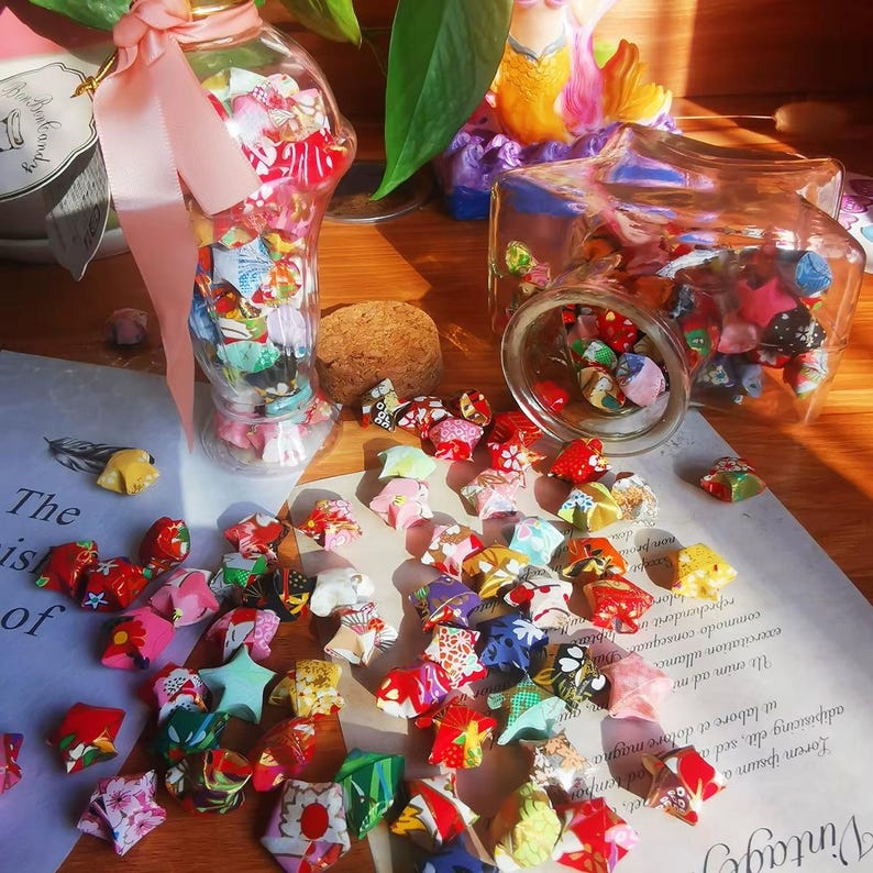 May include: A collection of colorful origami stars scattered on a wooden surface, with some inside glass jars. The stars are made from patterned paper in various colors, including red, pink, and green. A clear glass bottle is filled with origami stars and tied with a pink ribbon.
