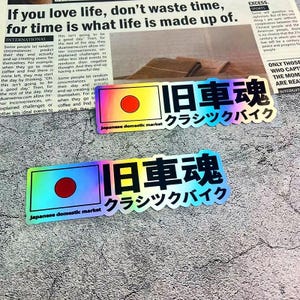 May include: Two holographic stickers with Japanese text and a rising sun design. The stickers read "Japanese domestic market" and "Classic Bike". The background is a newspaper.