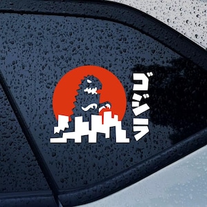 May include: White vinyl decal featuring a silhouette of a monster in front of an orange sun, with a cityscape below. Japanese text is to the right of the design. The decal is applied to a car window.